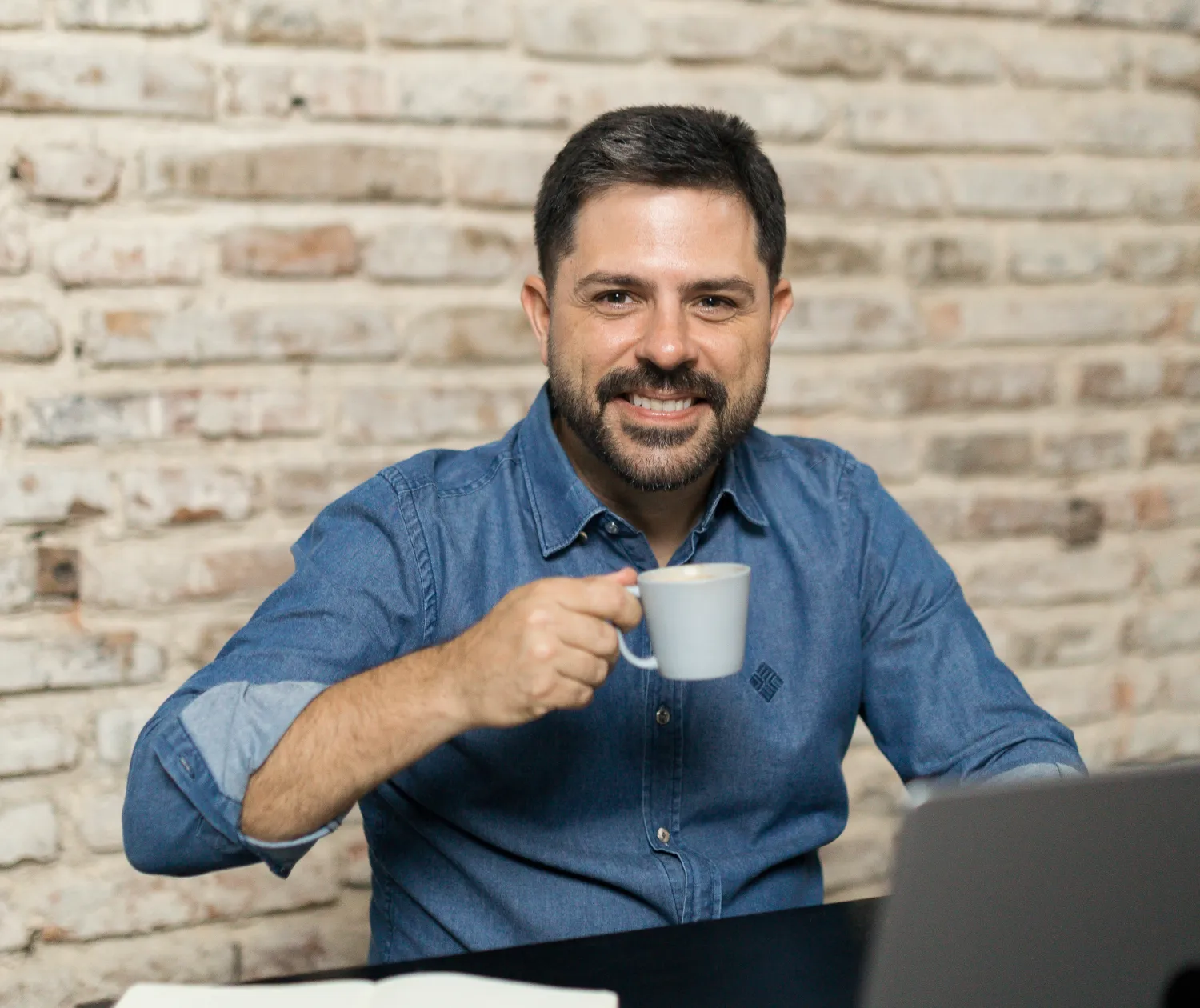 Tiago Asfora sorrindo, com uma xícara de café, na mesa de trabalho